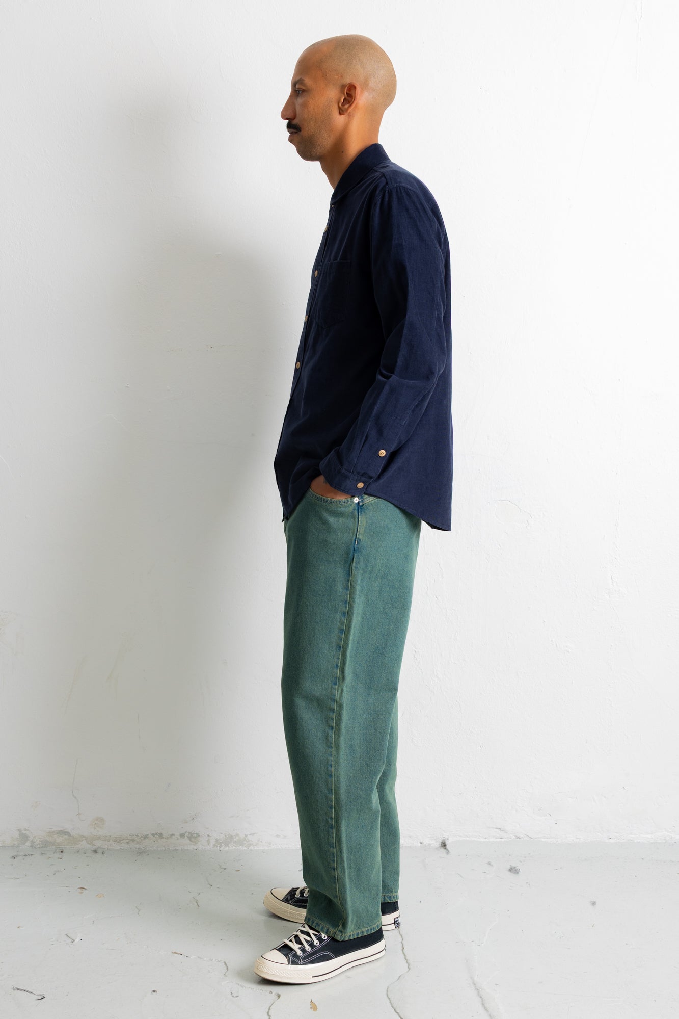 A man stands in profile wearing a dark navy long-sleeve button-up shirt, teal-green trousers, and black canvas sneakers against a plain white wall.