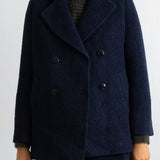 A person wearing a dark navy double-breasted textured coat over a dark ribbed turtleneck sweater and matching dark trousers, shown from the mouth down against a plain light background.