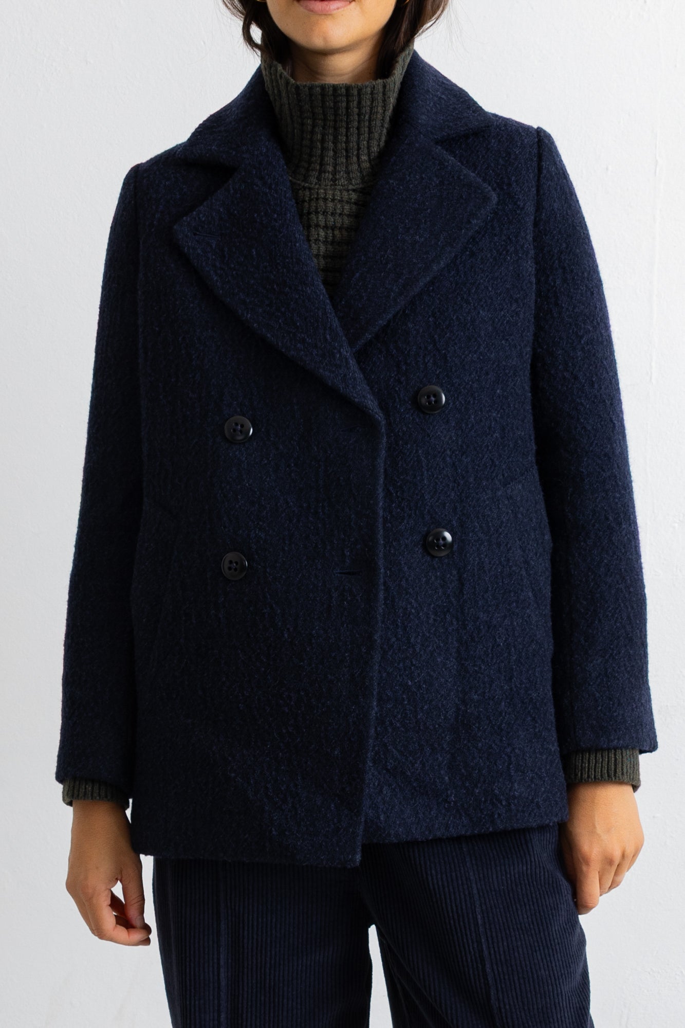 A person wearing a dark navy double-breasted textured coat over a dark ribbed turtleneck sweater and matching dark trousers, shown from the mouth down against a plain light background.