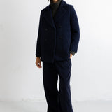 A model stands against a white studio wall wearing a dark navy double-breasted wool-blend coat with matching wide-leg trousers, a dark ribbed turtleneck, and black shoes.