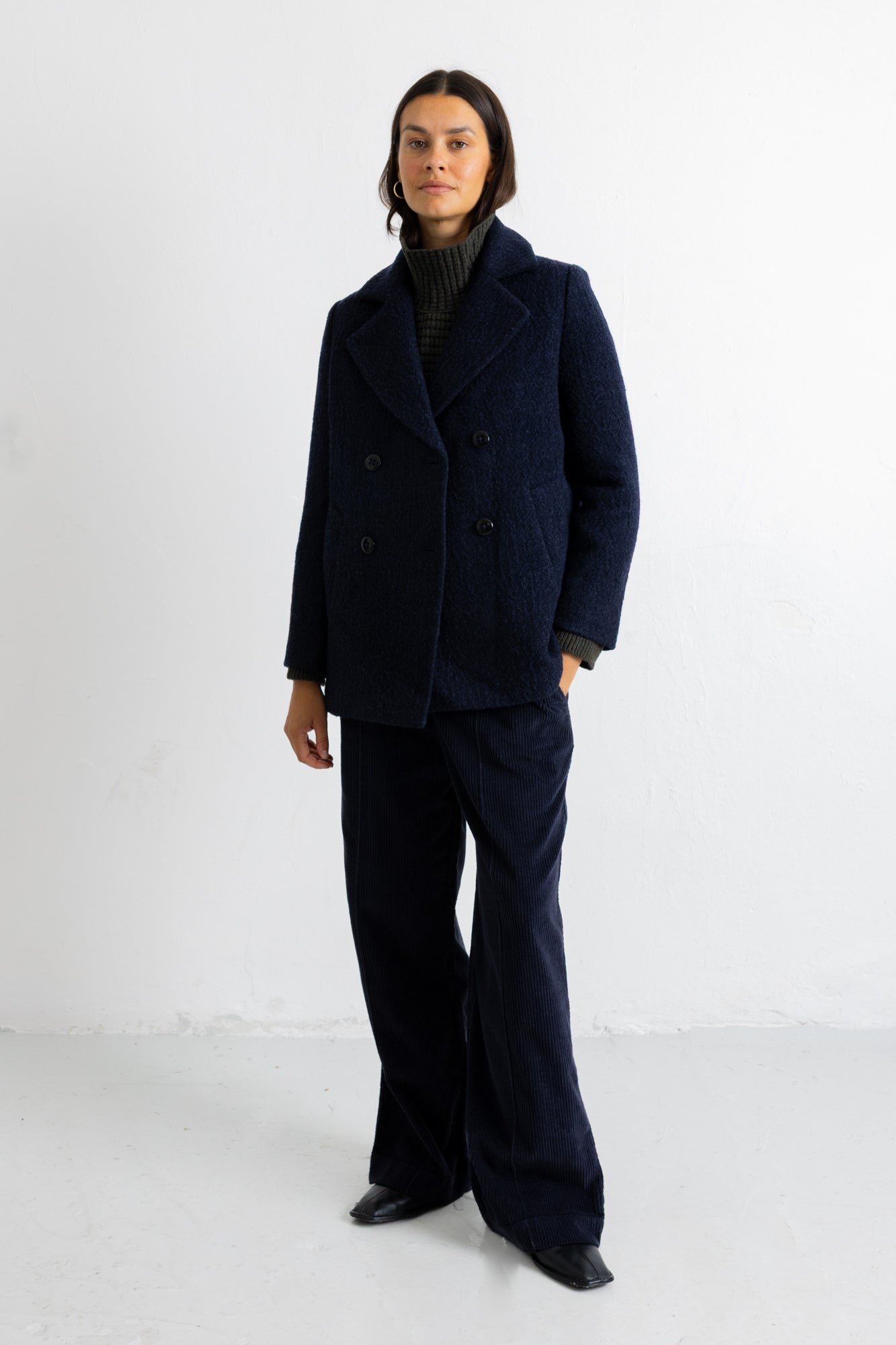 A model stands against a white studio wall wearing a dark navy double-breasted wool-blend coat with matching wide-leg trousers, a dark ribbed turtleneck, and black shoes.