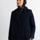 A woman wearing a dark navy blue double-breasted wool coat over a dark ribbed turtleneck sweater, standing against a plain white wall.