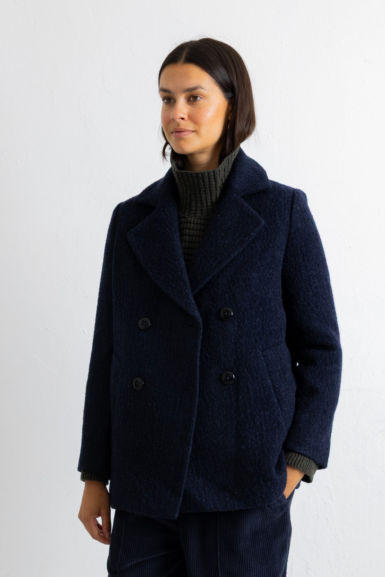 A woman wearing a dark navy blue double-breasted wool coat over a dark ribbed turtleneck sweater, standing against a plain white wall.