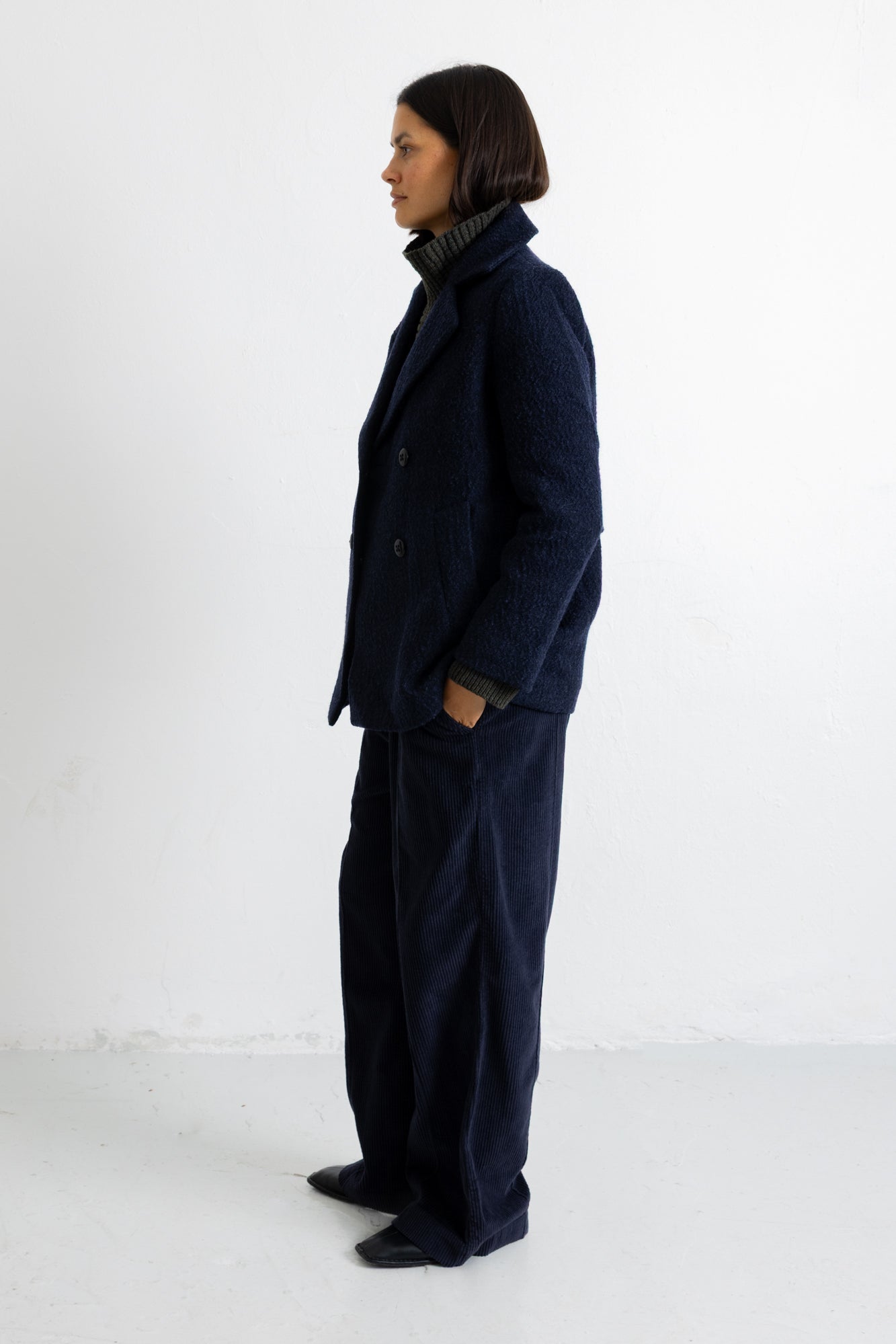 A model stands in profile wearing a dark navy blue textured jacket and matching wide-leg trousers, photographed against a plain white studio background.