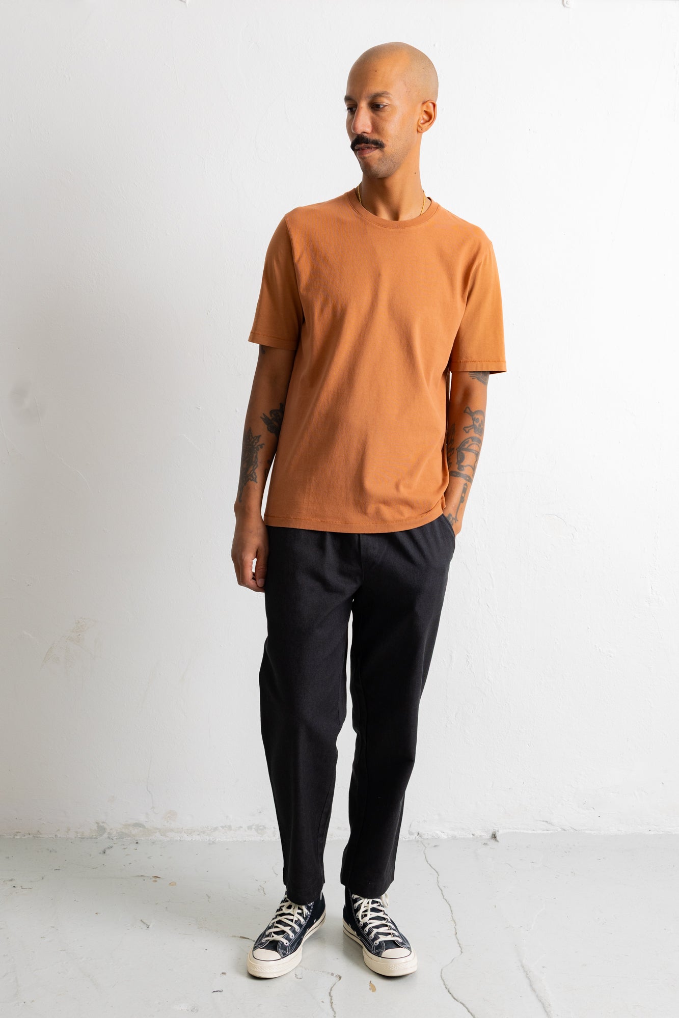 A man wearing a short-sleeve crewneck t-shirt in a muted orange, paired with black trousers and black-and-white sneakers, standing against a white wall.