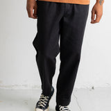 A person wearing loose black pants with an orange t-shirt and black low-top sneakers, standing against a white wall.