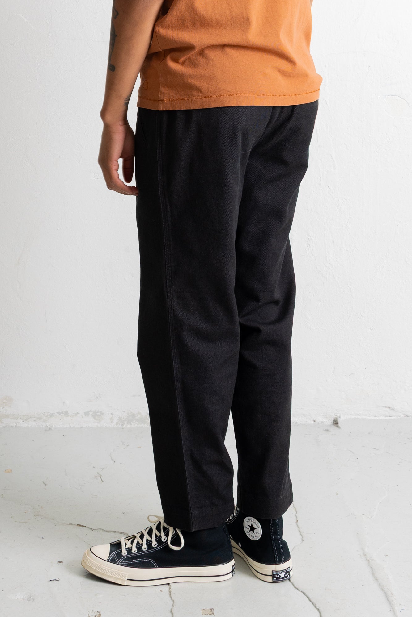 A person shown from the waist down wearing loose black pants, an orange t-shirt, and black Converse high-top sneakers against a white wall.