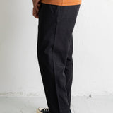 A person shown from the waist down wearing dark charcoal gray relaxed-fit pants, an orange t-shirt, and black canvas sneakers with white laces, standing against a white wall.