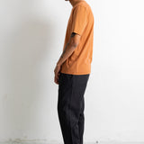 A man in profile wearing a short-sleeve crewneck t-shirt in a muted orange color, paired with dark charcoal pants and black canvas sneakers, standing against a white wall.