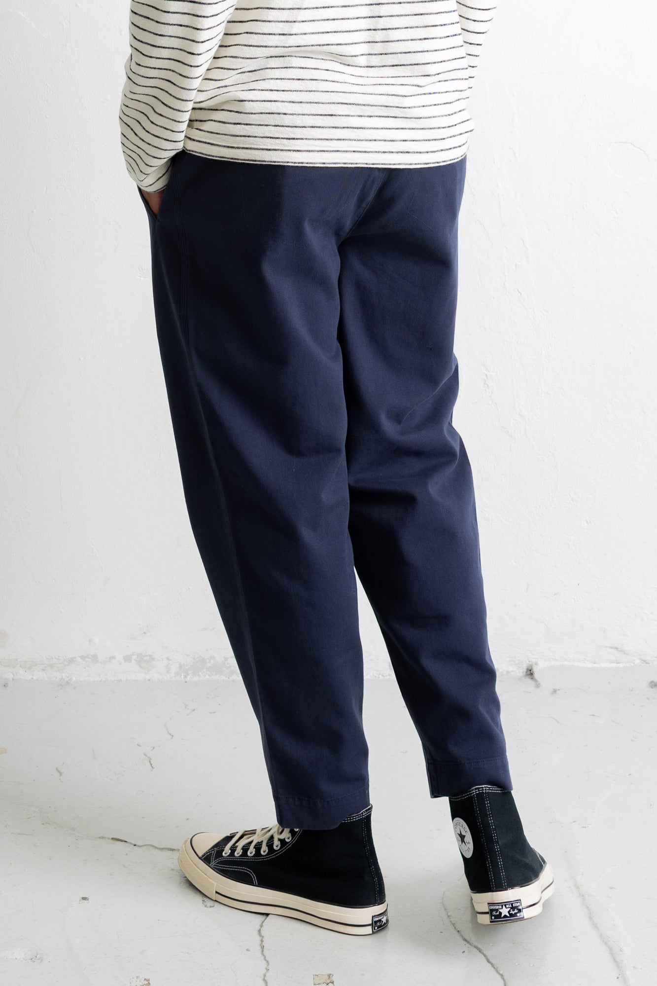 A person shown from the waist down wearing navy blue relaxed-fit trousers, a cream and navy striped long-sleeve shirt, and black high-top sneakers with white soles, standing against a white wall.