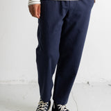 A person wearing navy blue relaxed-fit trousers, a cream and navy striped long-sleeve shirt, and black canvas low-top sneakers with white rubber soles, standing against a white wall.