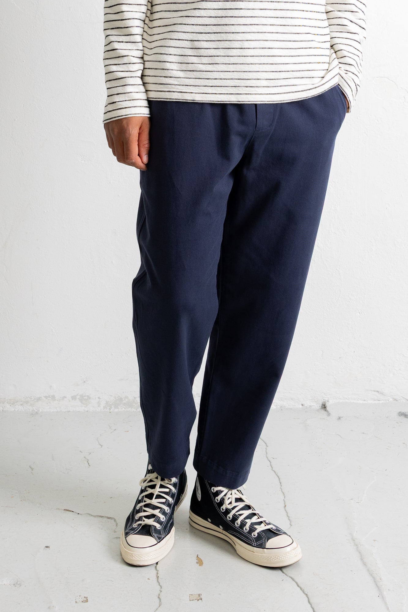 A person wearing navy blue relaxed-fit trousers, a cream and navy striped long-sleeve shirt, and black canvas low-top sneakers with white rubber soles, standing against a white wall.