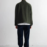 A person shown from the back wearing an olive green jacket, dark navy pants, a dark navy beanie, and black shoes, standing against a plain white wall.