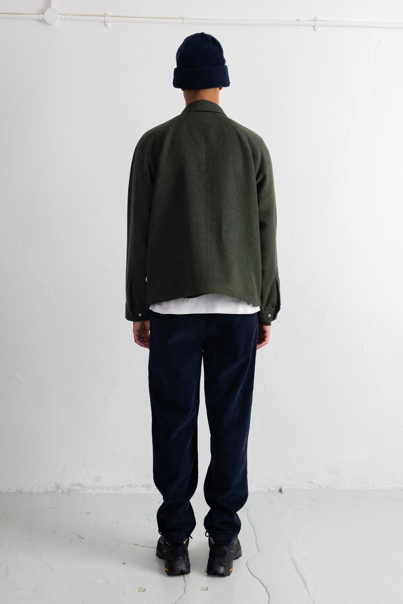 A person shown from the back wearing an olive green jacket, dark navy pants, a dark navy beanie, and black shoes, standing against a plain white wall.