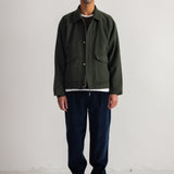 A man stands against a plain white studio wall wearing an olive green utility jacket over a white shirt, dark blue cords, black sneakers, and a dark navy beanie.