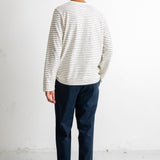 A man viewed from behind wearing a cream long-sleeve shirt with thin dark horizontal stripes, navy blue pants, and black canvas sneakers, standing against a white studio wall.