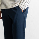 A close-up of a person wearing dark navy blue trousers and a cream-colored long-sleeve top with thin black horizontal stripes, standing against a white textured wall.