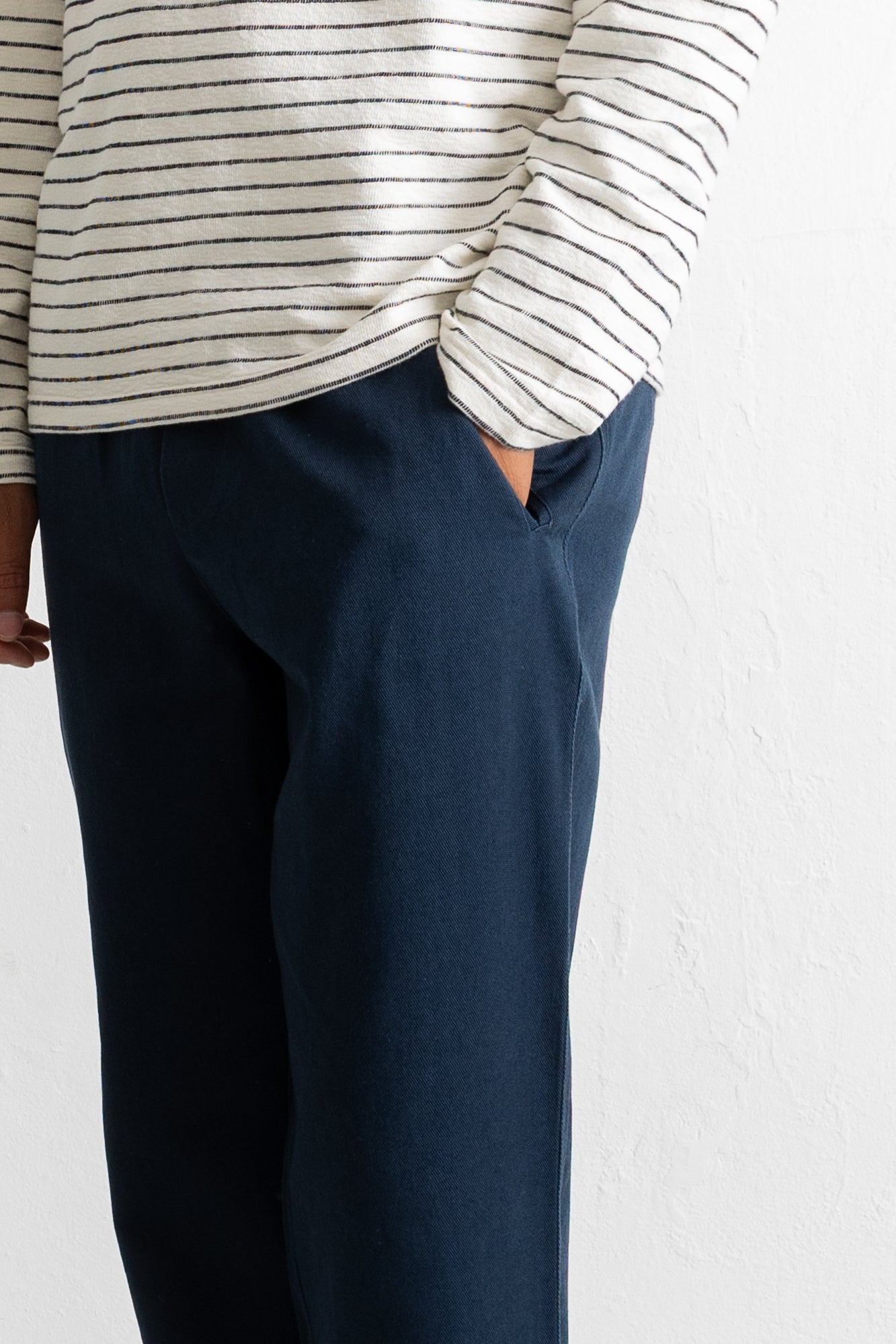 A close-up of a person wearing dark navy blue trousers and a cream-colored long-sleeve top with thin black horizontal stripes, standing against a white textured wall.