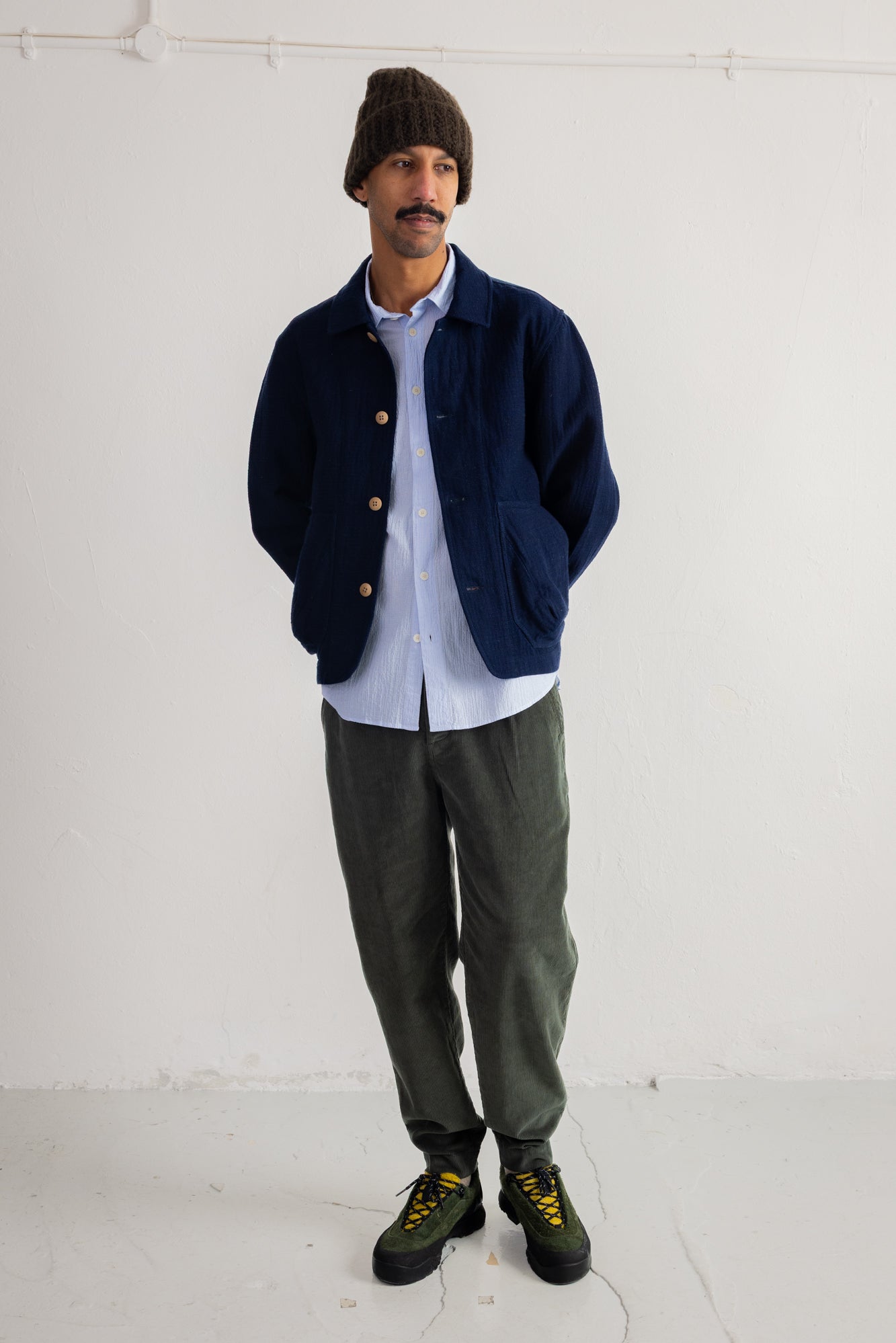 A man wearing a navy blue corduroy jacket over a light blue button-down shirt, paired with dark green trousers and black boots, standing against a plain white wall.