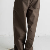 A person wearing loose brown pants with a white t-shirt and black canvas sneakers, shown from the waist down against a light gray wall and floor.