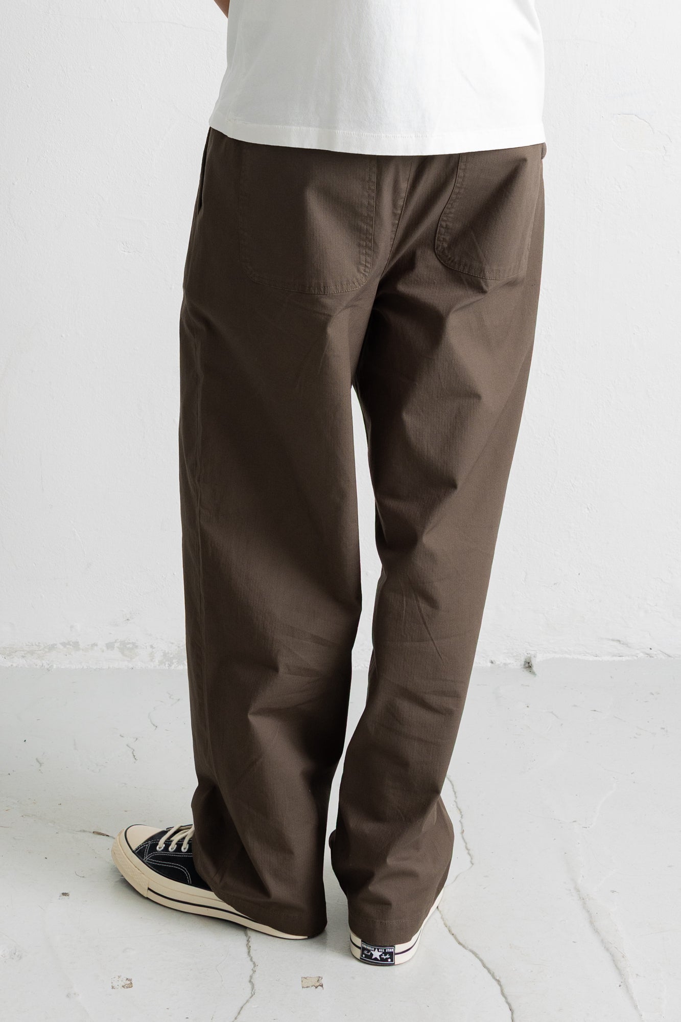 A person wearing loose brown pants with a white t-shirt and black canvas sneakers, shown from the waist down against a light gray wall and floor.