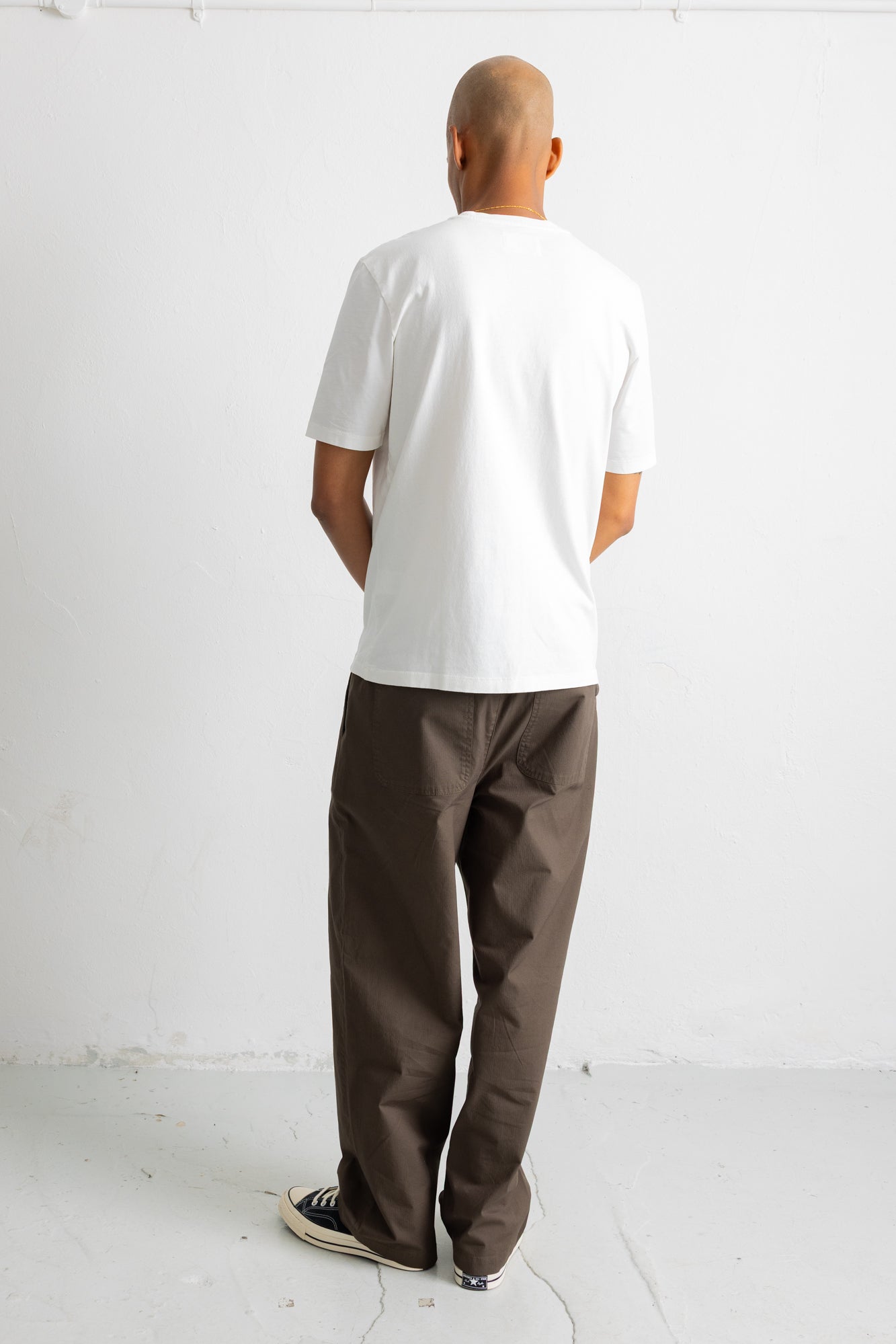 A man viewed from behind wearing a plain white short-sleeve t-shirt and dark brown relaxed-fit pants, standing against a white studio wall.