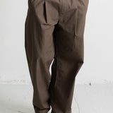 A person wearing loose, wide-leg brown pants with a white t-shirt and black canvas sneakers, standing against a light gray wall.