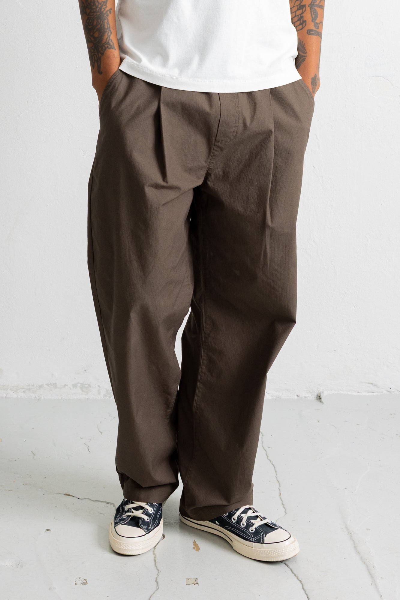 A person wearing loose, wide-leg brown pants with a white t-shirt and black canvas sneakers, standing against a light gray wall.