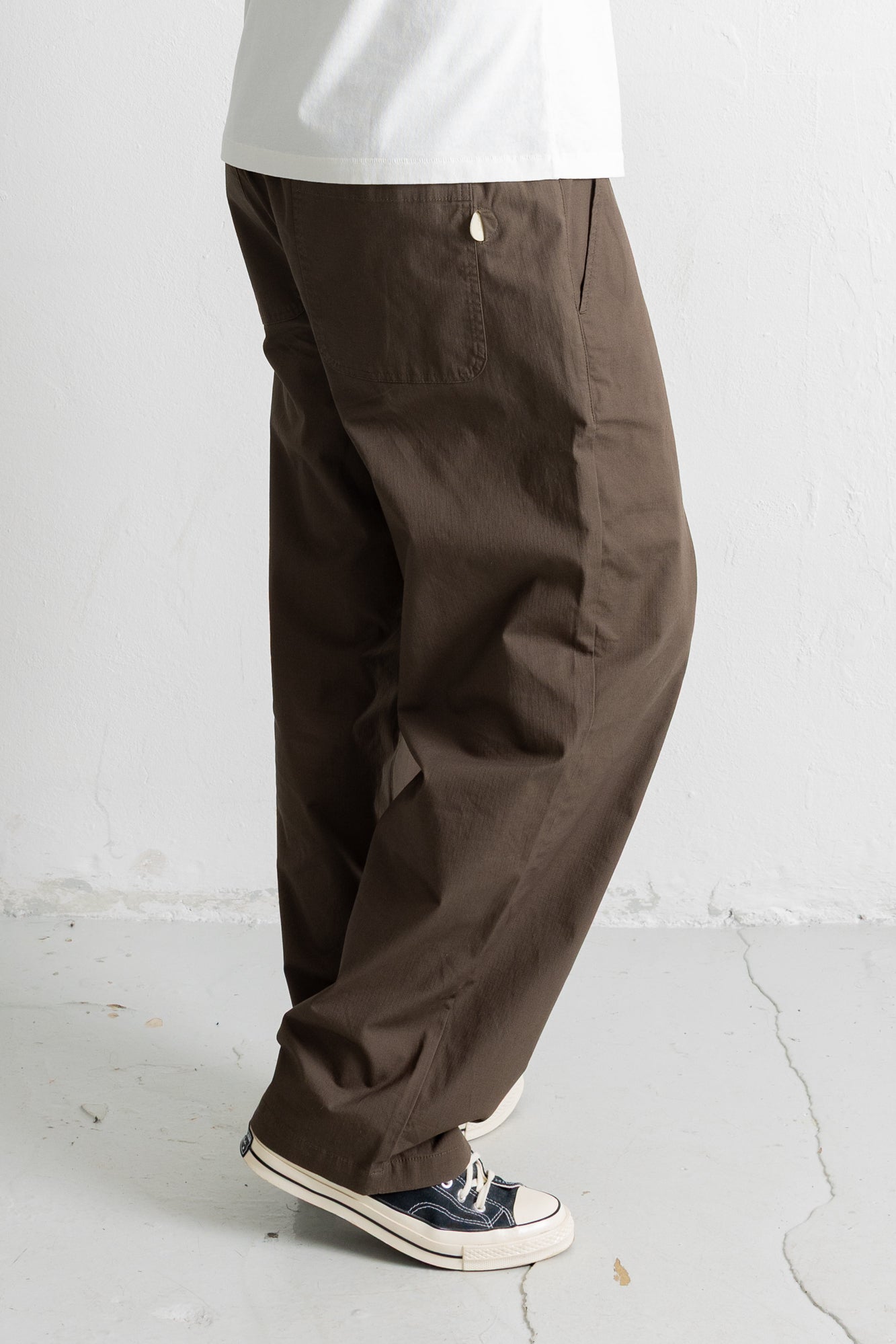 A person wearing loose, wide-leg brown pants with a white t-shirt and navy low-top sneakers, shown from the waist down against a light gray wall and floor.
