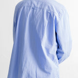 A man viewed from the back wearing a light blue long-sleeve button-up shirt and dark blue jeans, against a plain white wall.