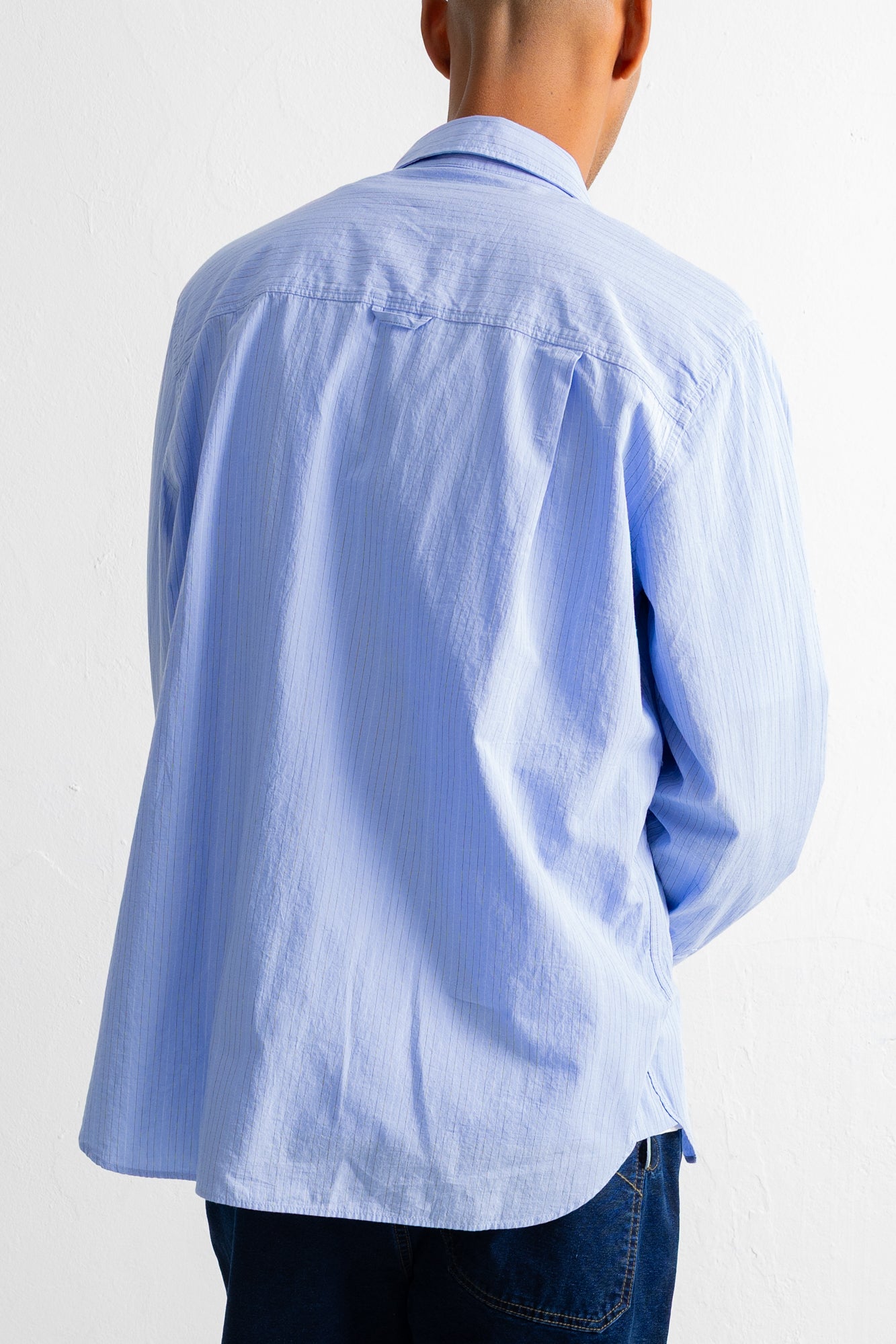 A man viewed from the back wearing a light blue long-sleeve button-up shirt and dark blue jeans, against a plain white wall.
