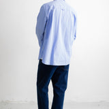 A man viewed from behind wearing a light blue long-sleeve button-down shirt, dark navy pants, and black canvas sneakers, standing against a plain white wall.