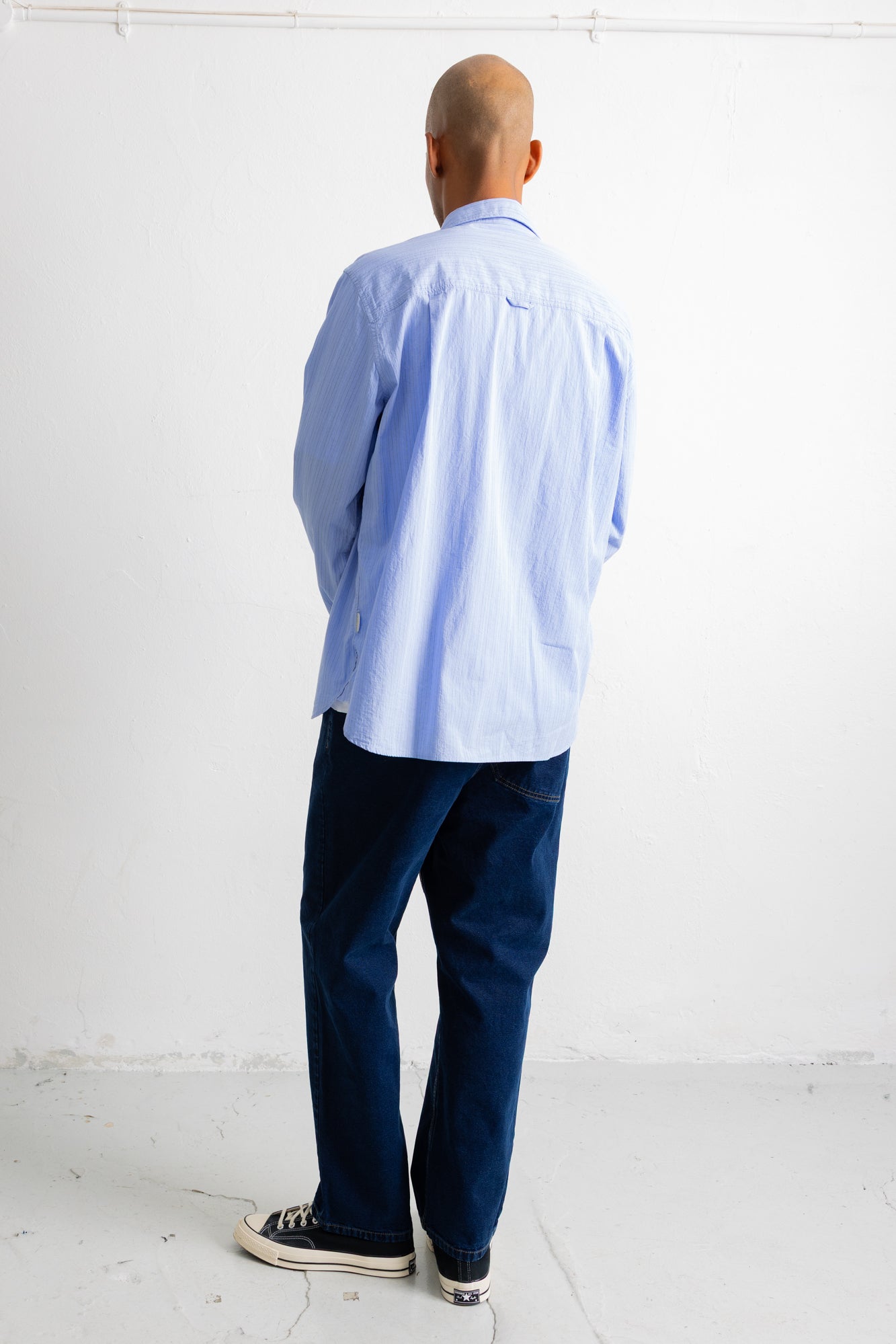 A man viewed from behind wearing a light blue long-sleeve button-down shirt, dark navy pants, and black canvas sneakers, standing against a plain white wall.