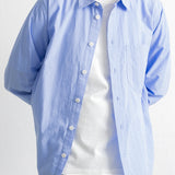 A man wearing a light blue long-sleeve button-down shirt over a white t-shirt and dark blue jeans, shown from the neck down against a plain white background.