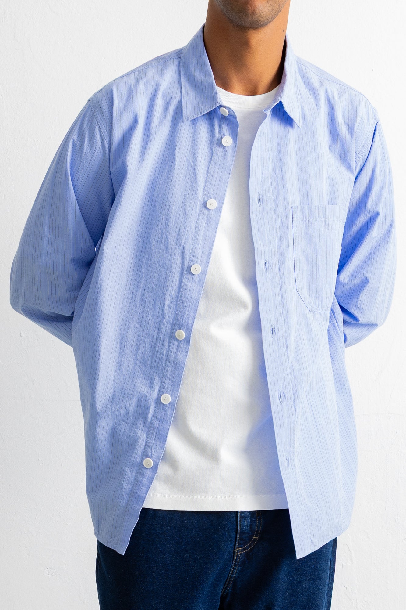 A man wearing a light blue long-sleeve button-down shirt over a white t-shirt and dark blue jeans, shown from the neck down against a plain white background.