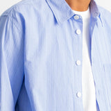 A close-up of a man wearing a light blue and white vertically striped button-down shirt over a white t-shirt, cropped from the chin down.