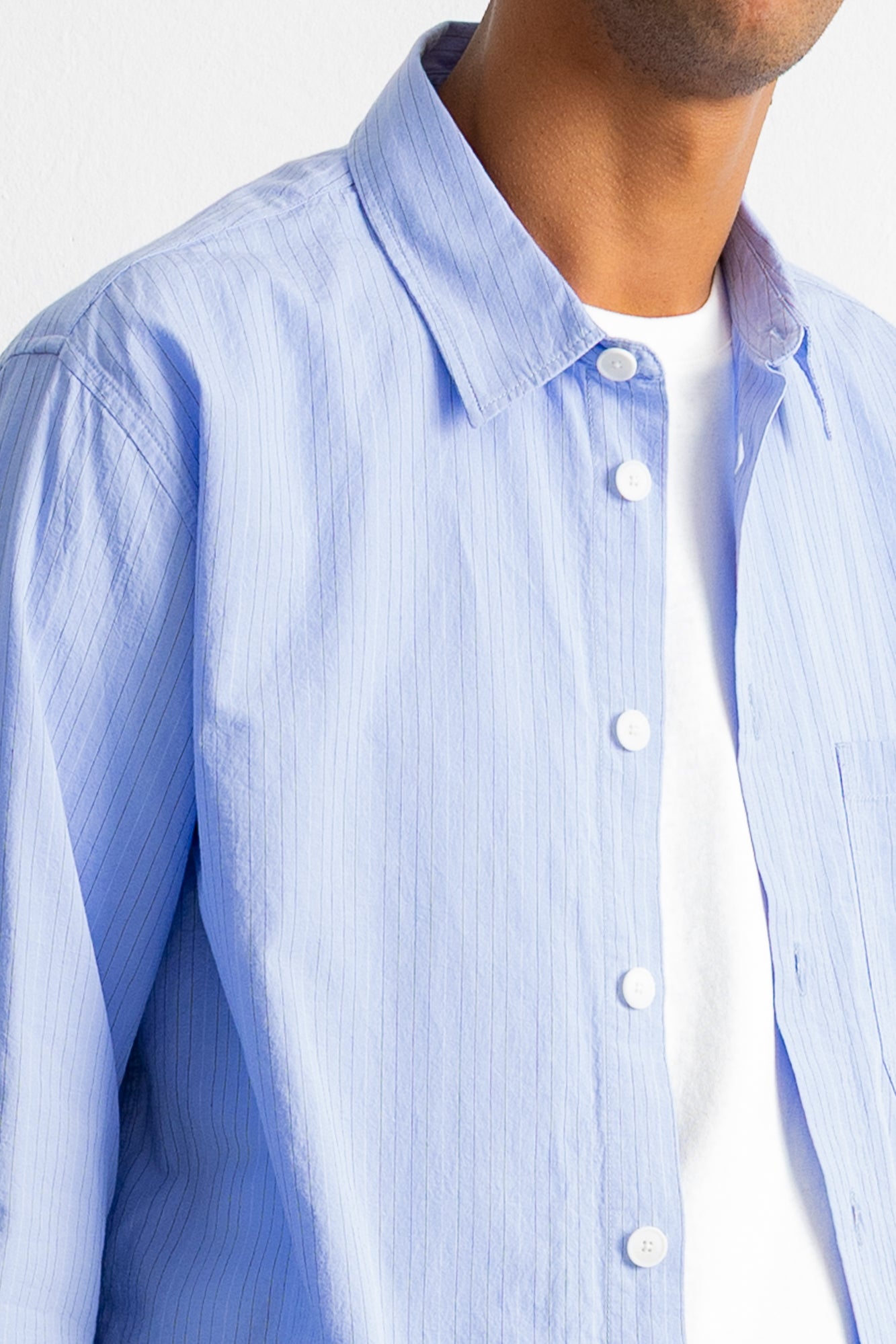 A close-up of a man wearing a light blue and white vertically striped button-down shirt over a white t-shirt, cropped from the chin down.