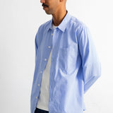 A man wearing a light blue long-sleeve button-down shirt over a white t-shirt and dark blue jeans, standing against a plain white wall.