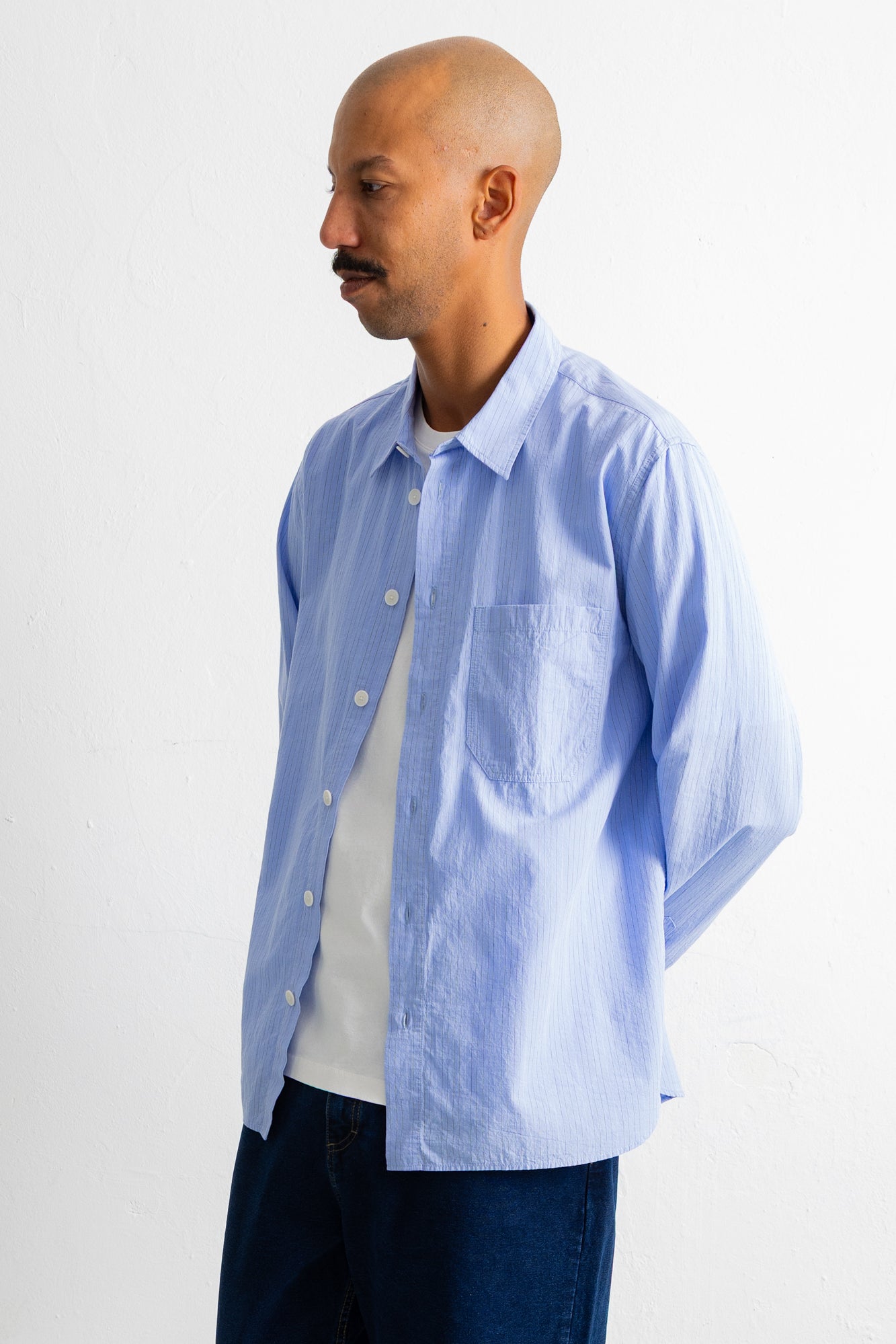 A man wearing a light blue long-sleeve button-down shirt over a white t-shirt and dark blue jeans, standing against a plain white wall.