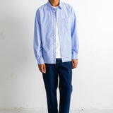 A man wearing a light blue long-sleeve button-down shirt over a white t-shirt, paired with dark blue jeans and black-and-white sneakers, standing against a white wall.