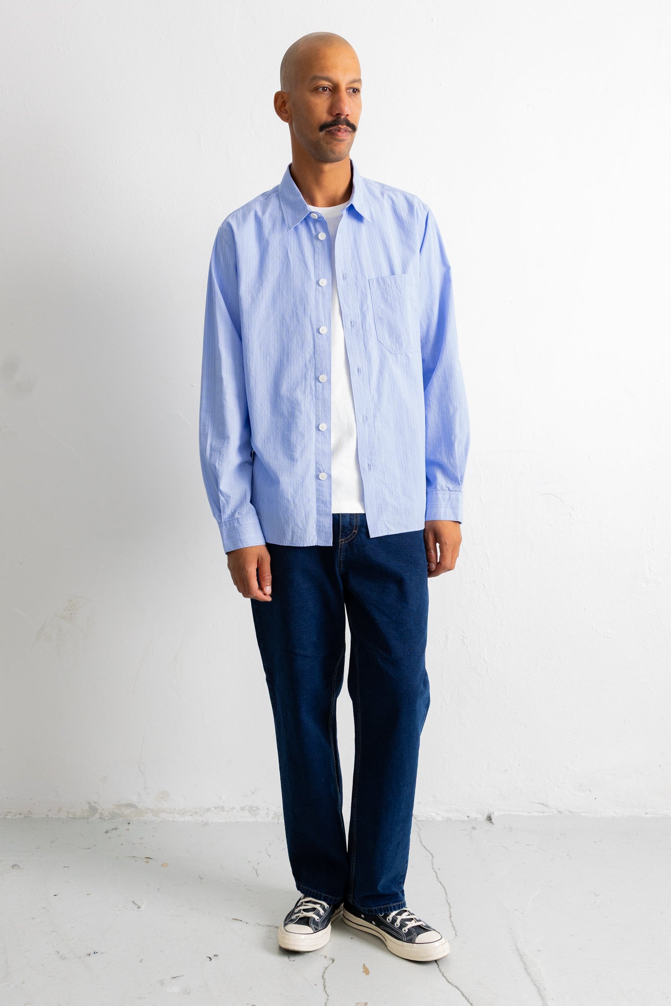 A man wearing a light blue long-sleeve button-down shirt over a white t-shirt, paired with dark blue jeans and black-and-white sneakers, standing against a white wall.