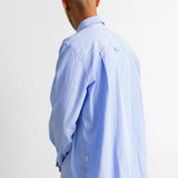 A man viewed from behind wearing a light blue long-sleeve button-down shirt and dark blue jeans against a plain white wall.