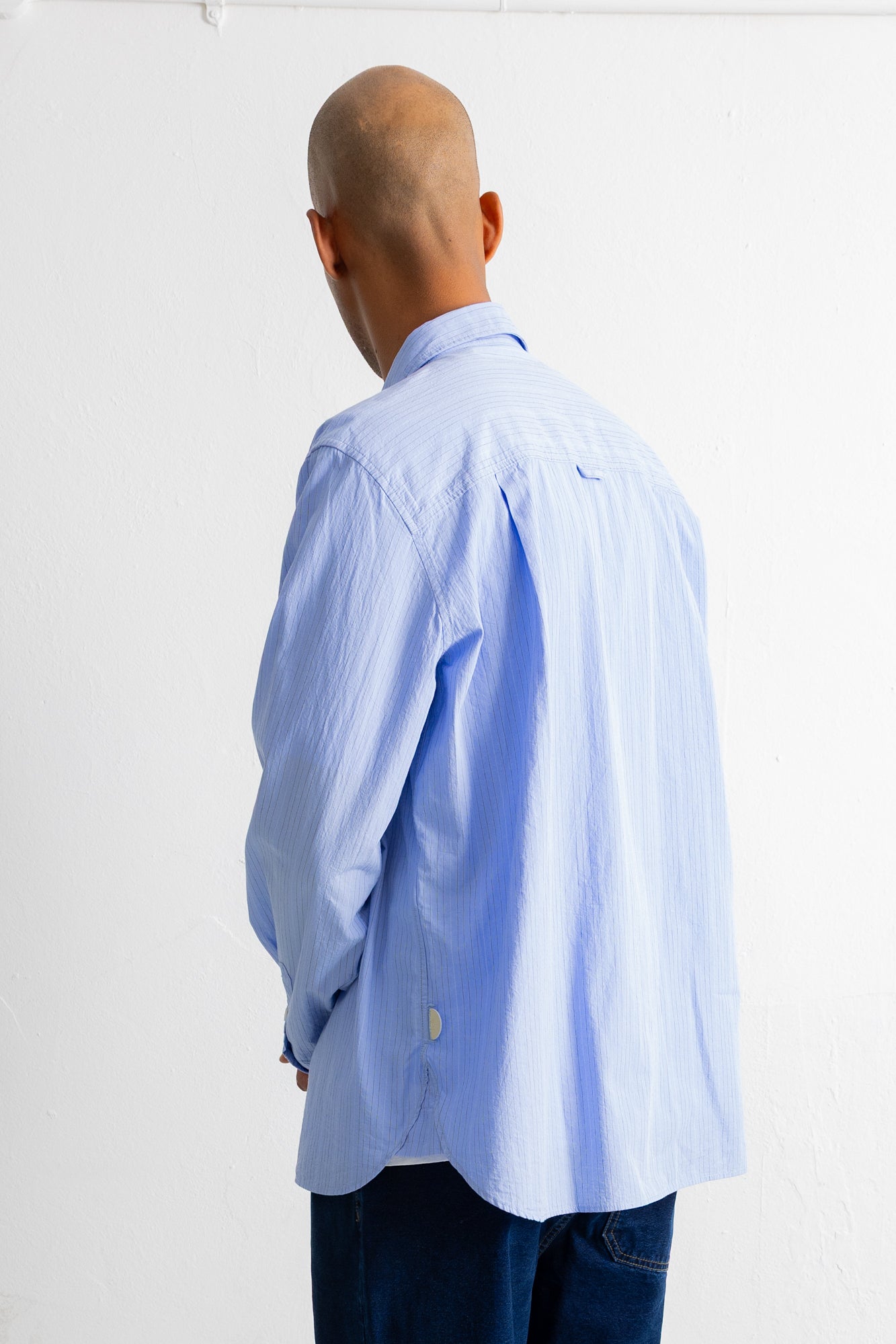 A man viewed from behind wearing a light blue long-sleeve button-down shirt and dark blue jeans against a plain white wall.