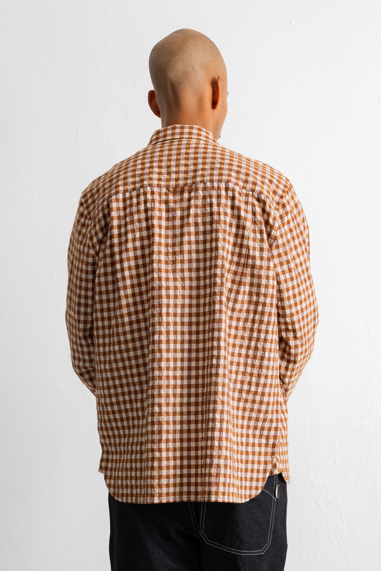 A man viewed from behind wearing a brown and white gingham check long-sleeve shirt and dark jeans against a plain white background.