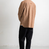 A man viewed from behind wearing a brown and beige checkered long-sleeve shirt, black jeans, and black canvas sneakers, standing against a plain white wall.