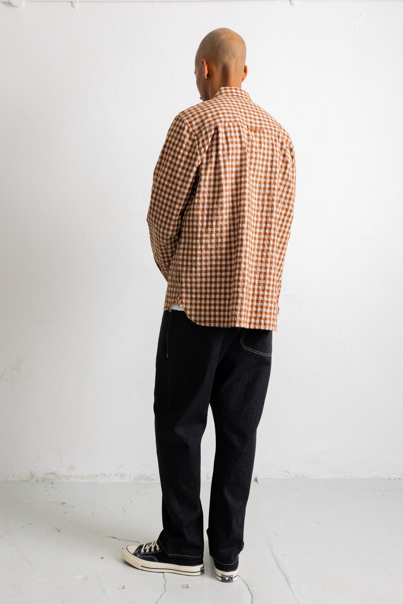 A man viewed from behind wearing a brown and beige checkered long-sleeve shirt, black jeans, and black canvas sneakers, standing against a plain white wall.