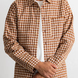 A man wearing a brown and beige gingham check long-sleeve button-up shirt over a white t-shirt and dark jeans, shown from the chin down against a plain white background.