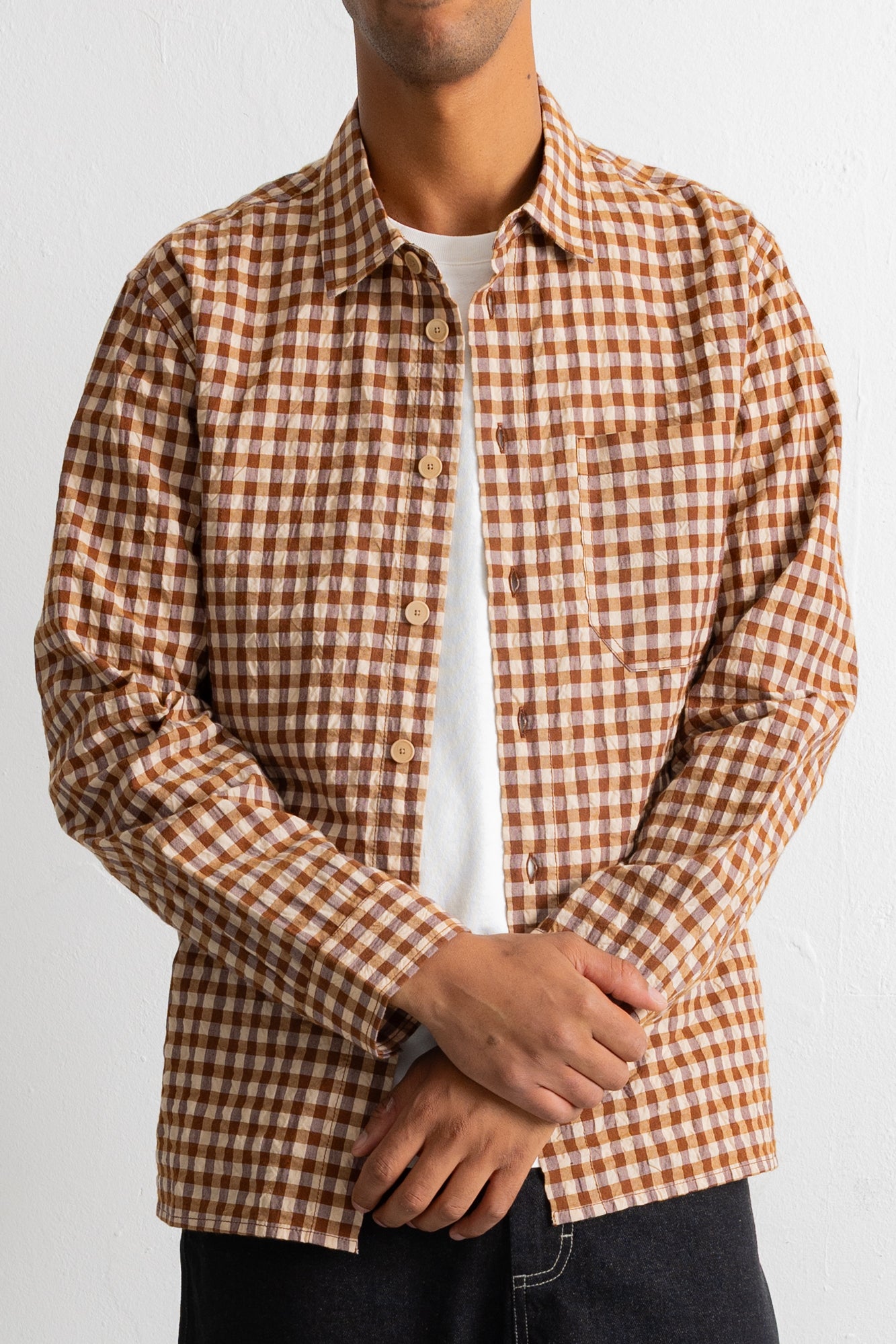 A man wearing a brown and beige gingham check long-sleeve button-up shirt over a white t-shirt and dark jeans, shown from the chin down against a plain white background.