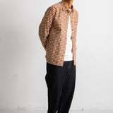 A man wearing a brown and beige checkered button-up shirt over a white t-shirt, black pants, and black canvas sneakers, standing against a white wall.