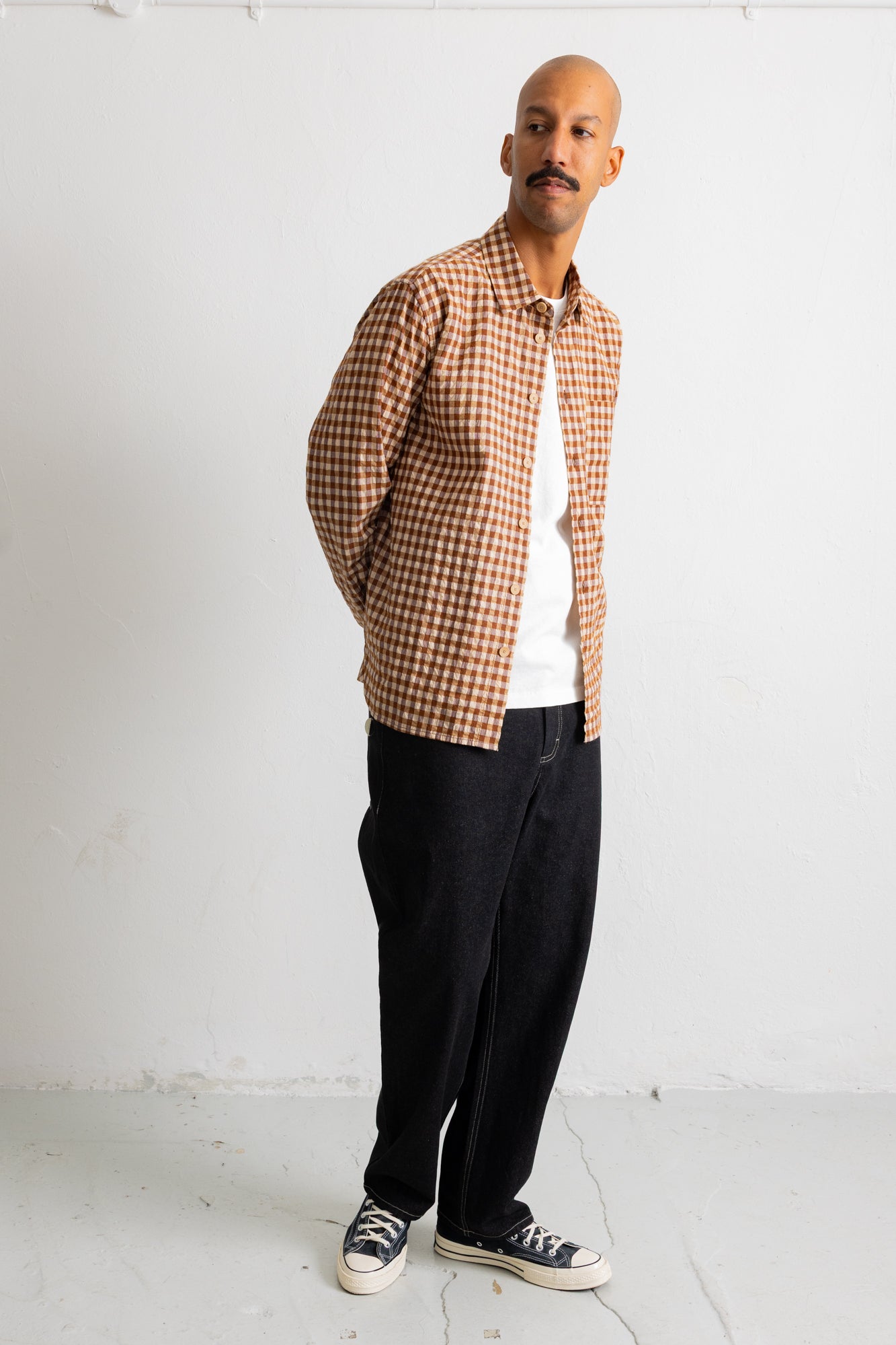 A man wearing a brown and beige checkered button-up shirt over a white t-shirt, black pants, and black canvas sneakers, standing against a white wall.